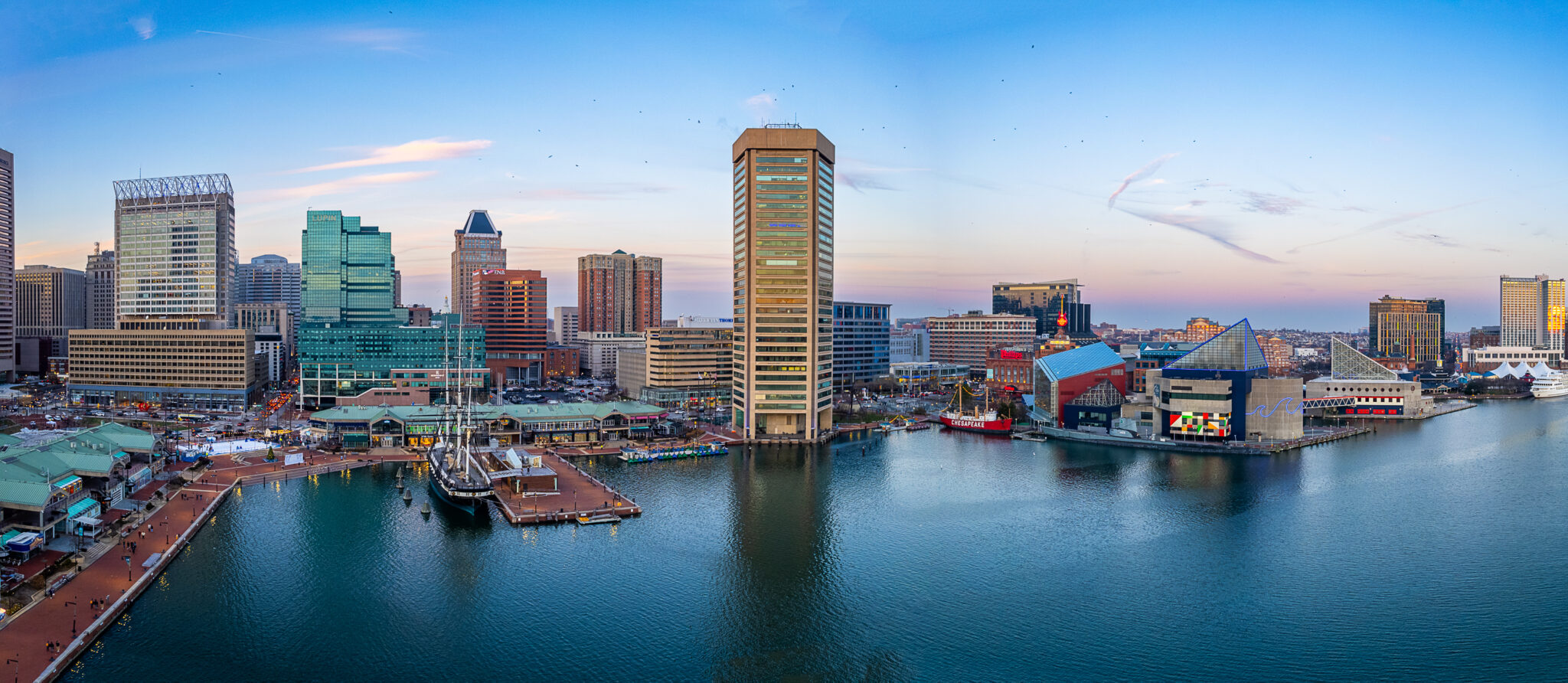 A panoramic view of Baltimore's Inner Harbor at sunset, showcasing a mix of modern and historic buildings along the waterfront.